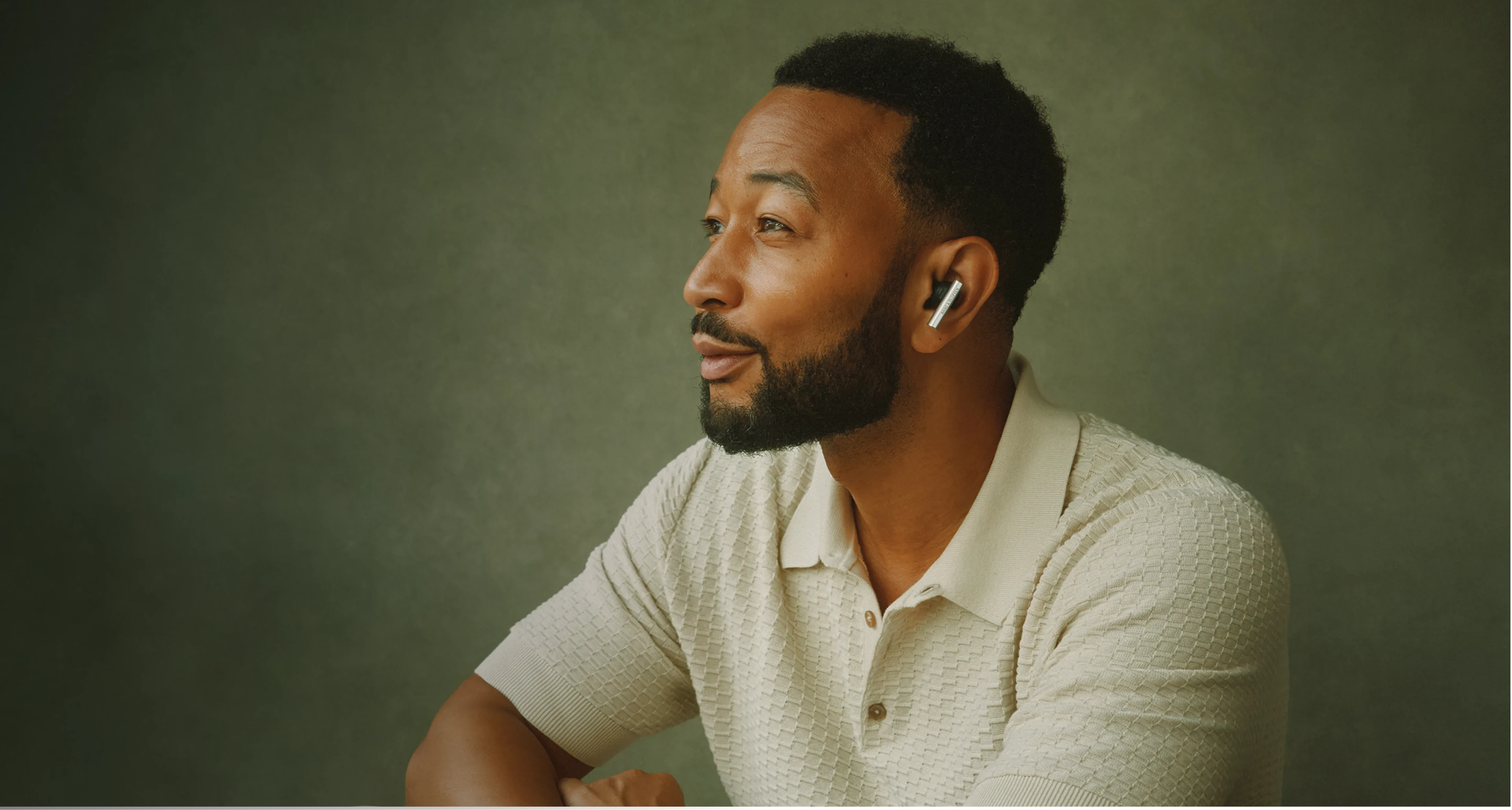 Our Partners wearing a cream knit polo shirt and SuperHoodie gazes thoughtfully to the side against a muted green background.