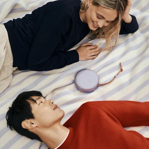 A couple lying face-to-face on a picnic mat with a Super Easy Apparel Manufacturing Studio Line A1 in Lavender placed between them, sharing a relaxed moment through music.